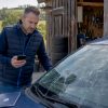 Un homme examine de près un impact sur le pare-brise de sa voiture, à l’extérieur.